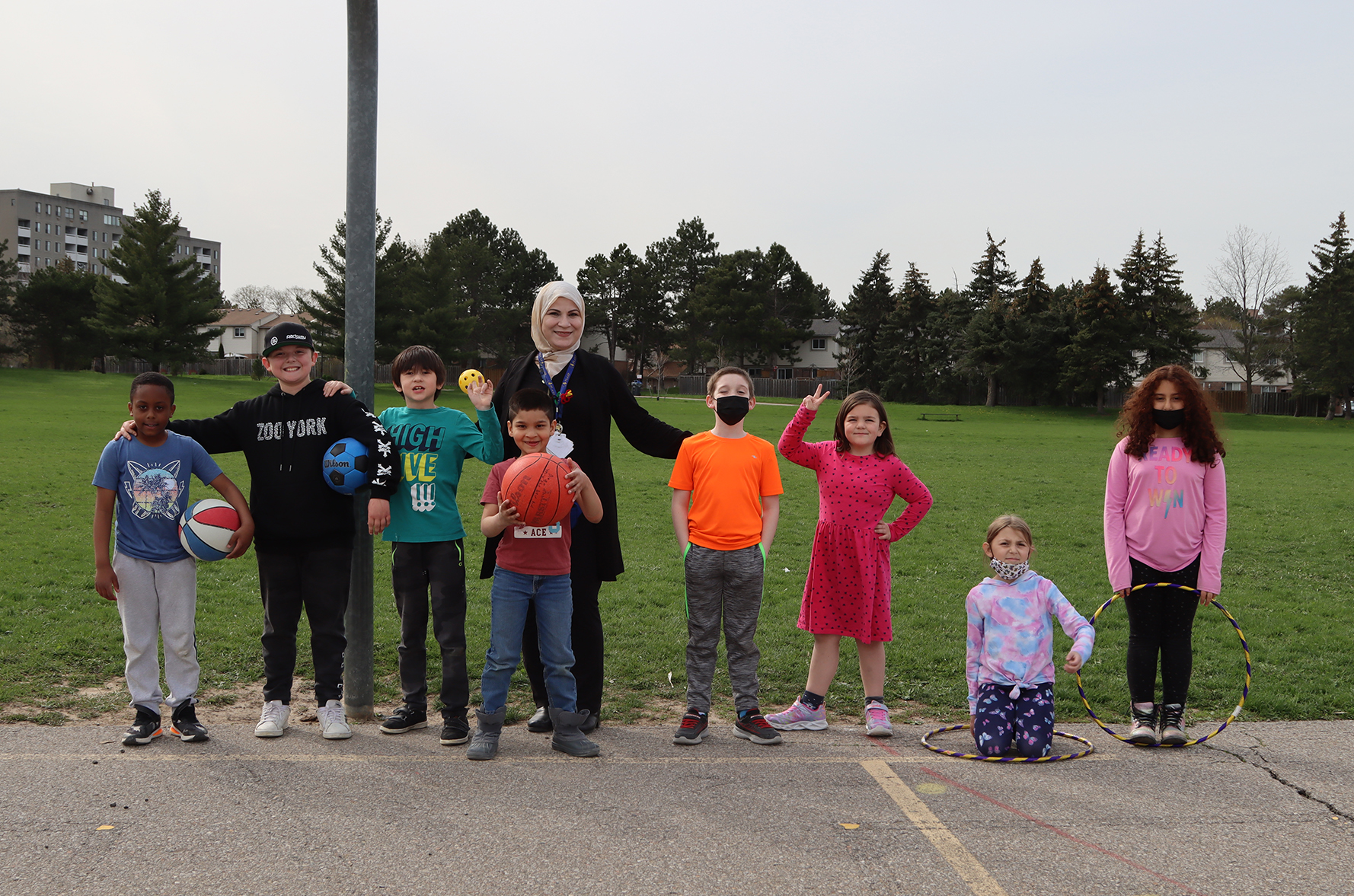 A PLASP Early Childhood Educator stands in a line with a group of children in program, some with a basketball and hula hoops